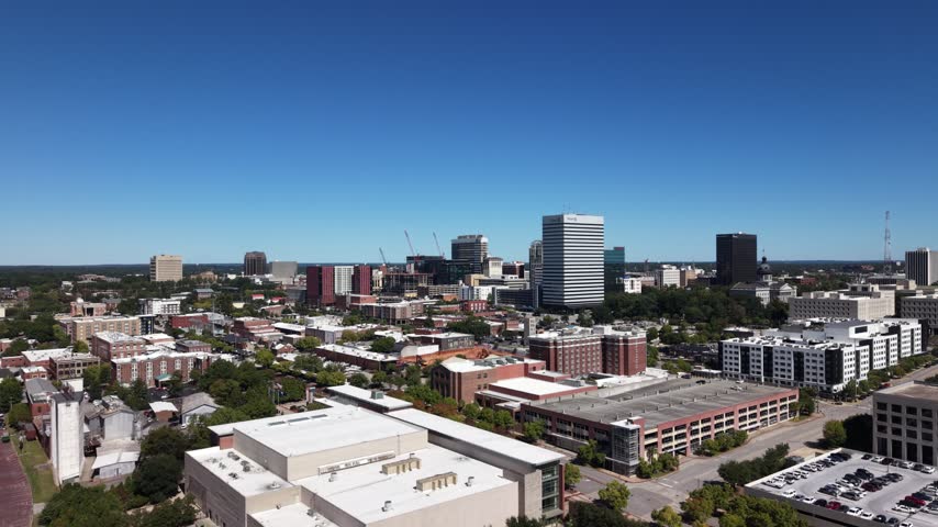 Drone aerial of the Columbia, South Carolina skyline with downtown towers rising over tree-lined streets and neighborhoods, traffic moving along the avenues below under a vast clear blue sky