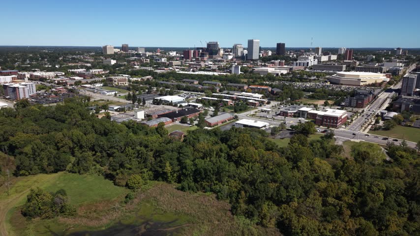 Drone aerial of the Columbia, South Carolina skyline with downtown towers rising over tree-lined streets and neighborhoods, traffic moving along the avenues below under a vast clear blue sky