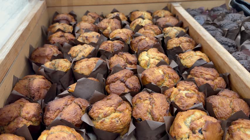 Rows of golden berry muffins in brown paper liners fill a large wooden box counter display, showcasing fresh sweet baked cupcakes for pastry cafe retail.