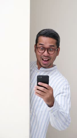 Adult Asian man peeking from behind white wall holding mobile phone with cheering expression