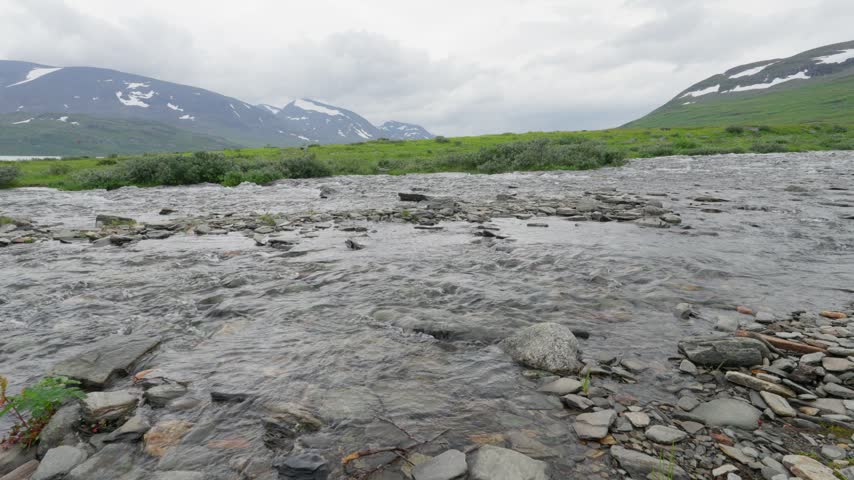 A lone hiker navigates a rocky river crossing on the Kungsleden trail in Sweden. The subarctic landscape features grassy hills and distant mountains, highlighting the remote nature adventure.