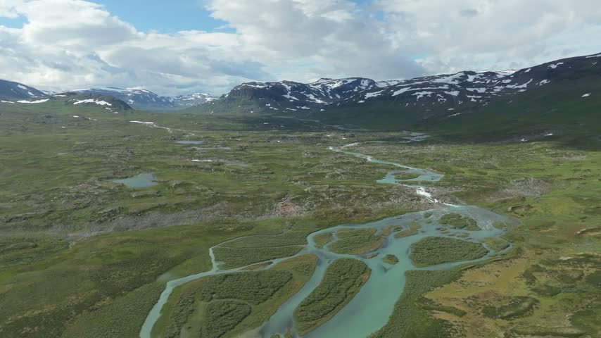 An aerial pull back drone shot revealing the Kungsleden hiking trail in a vast river valley in Sweden. Snow-capped mountains frame a lush, green landscape with a braided river system.