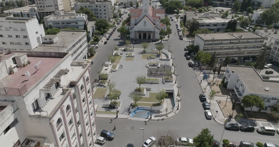 An aerial view changes perspective over an urban square with geometric planters, play areas, trees and surrounding streets. Filmed in Tangier Morocco.
