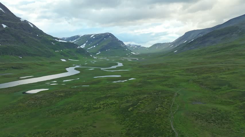 Aerial pull back drone shot revealing a vast, lush mountain valley along the Kungsleden trail in Sweden. The scene features a meandering river, small lakes, and snow-capped peaks under a cloudy sky.