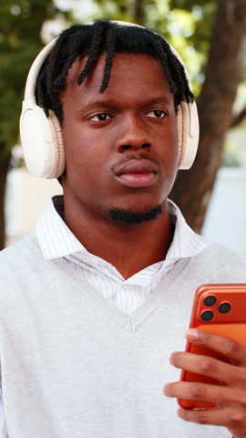 African American young man smiling with headphones walking in city street and looking at camera in sunny park. Black guy tourist enjoying music outdoors with smartphone in hand during calm park break