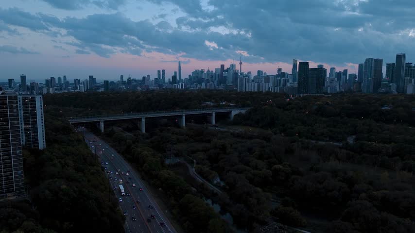Aerial editorial view of the Toronto skyline at dusk. The CN Tower and downtown high-rises stand against a pink and blue twilight sky, with a lit highway, long-exposure car light trails, a concrete bridge and dark autumn ravine forest below.