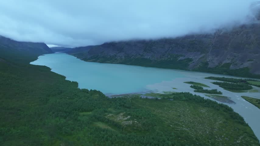 Aerial pan right over a stunning mountain lake and river in a green valley under a cloudy sky in Sweden, part of the Kungsleden hiking trail, surrounded by rocky mountains and lush forest.