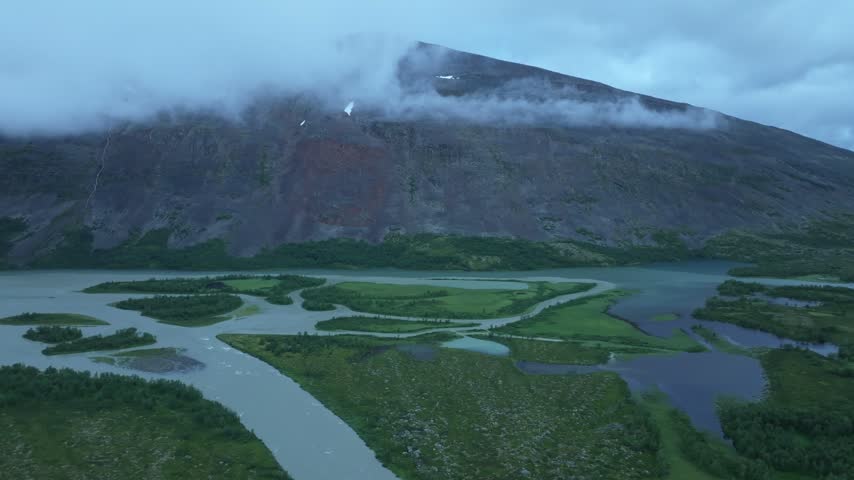 Aerial drone pan right across a stunning mountain landscape with a braided river, mountain tarns, and lush forest under an overcast sky. Close to the Kungsleden hiking trail.