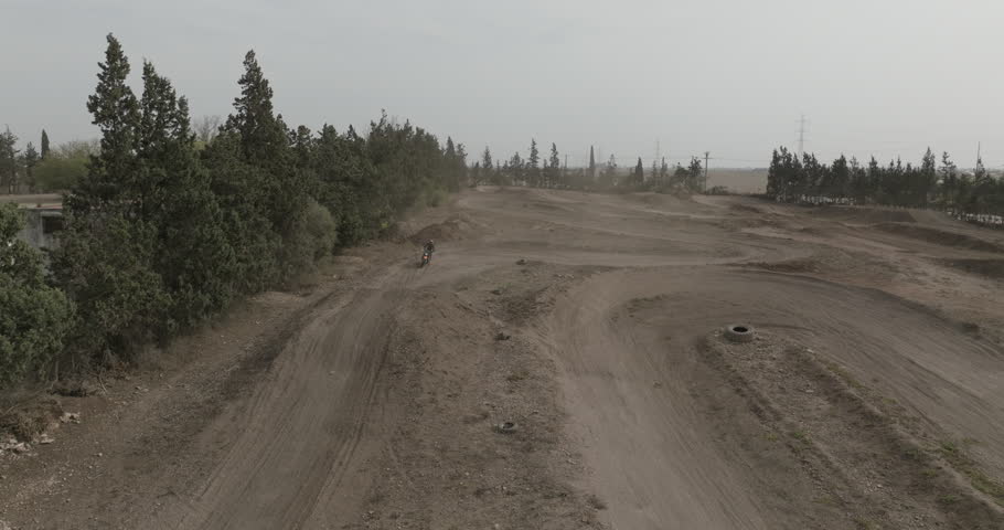 An aerial view follows a motocross rider approaching along a dirt course, crossing jumps and continuing after landing. Trees, tire markers and raised sections of track surround the rider.