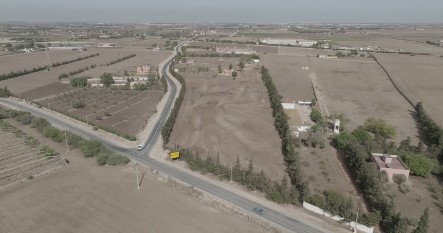 An aerial view moves from a rural road junction and nearby fields toward an overhead view of a dirt course. Trees border curved tracks and visible obstacles.