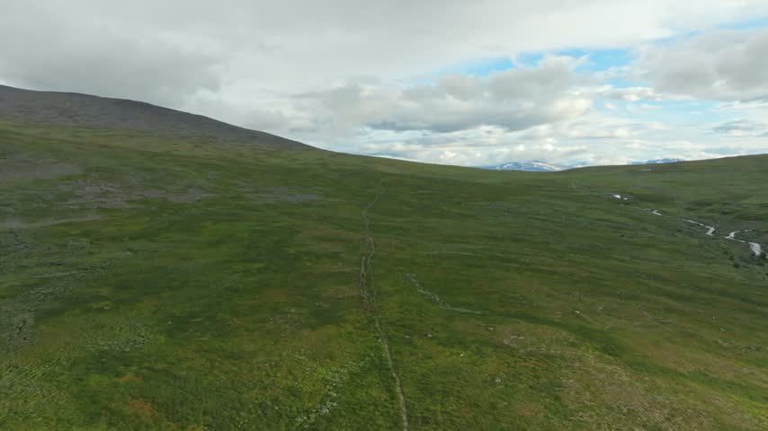 Aerial push in shot over the Kungsleden hiking trail in Sweden. The drone captures the vast, green tundra landscape with distant mountain ranges and a winding stream under a cloudy sky.