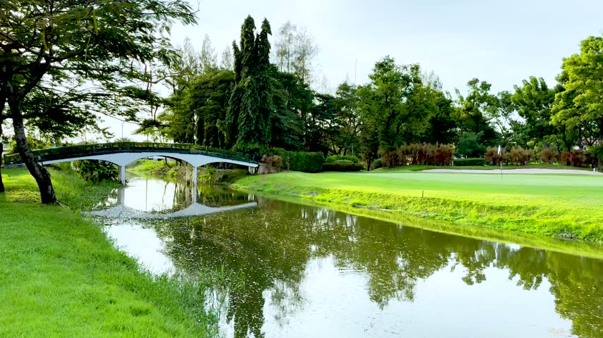 Peaceful park landscape featuring arched footbridge over water canal leading towards lush green golf course putting area.