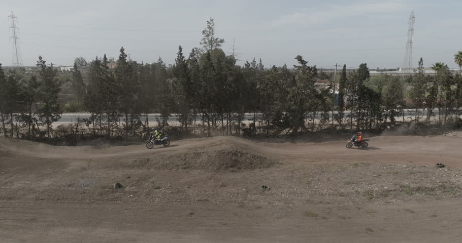 An aerial view follows several motocross riders moving over dirt mounds and track sections. A group of people and motorcycles stands near the center of the course as the camera pulls back to a wider view.