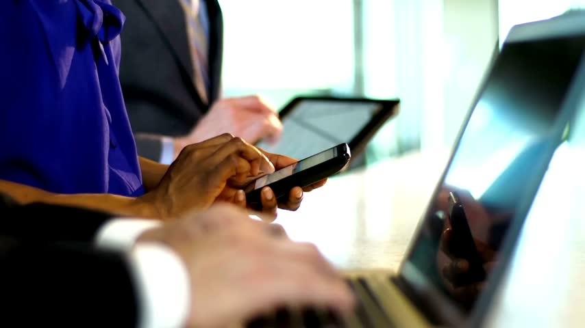 Close-up side view of corporate professionals using a laptop, smartphone, and digital tablet simultaneously in a modern office setup.