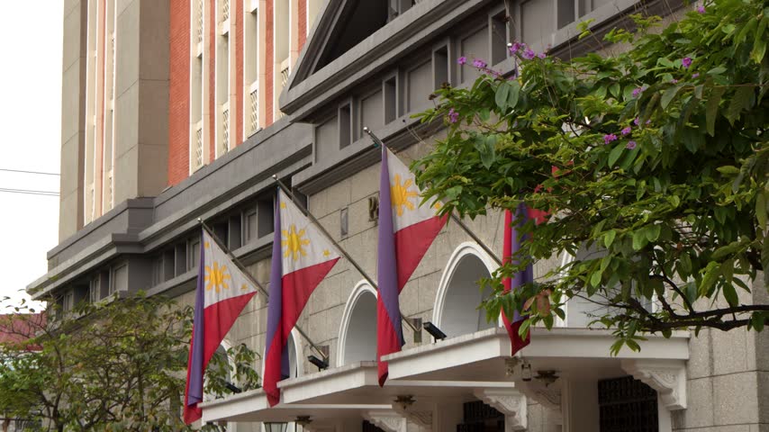 Rows of Philippines national flags attached to stone building exterior, waving softly in breeze during cloudy afternoon in Manila.
