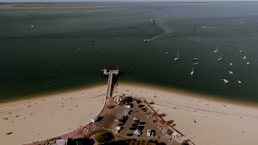 Jetee Thiers at Arcachon in Gironde, France, a jetty running out from the beach next to a vintage carousel and a seafront car park, with boats crossing the bay.