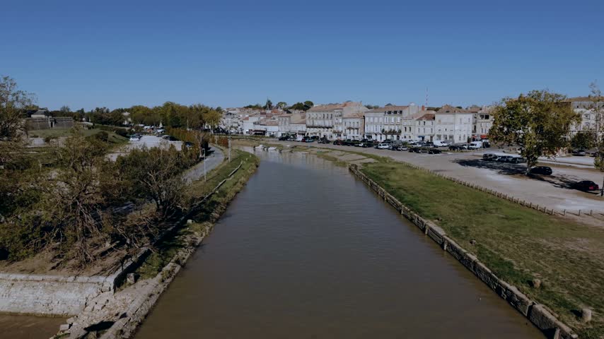 Tidal channel of the port of Blaye in Gironde, France, running between grassy embankments toward stone townhouses and parked cars on the waterfront.