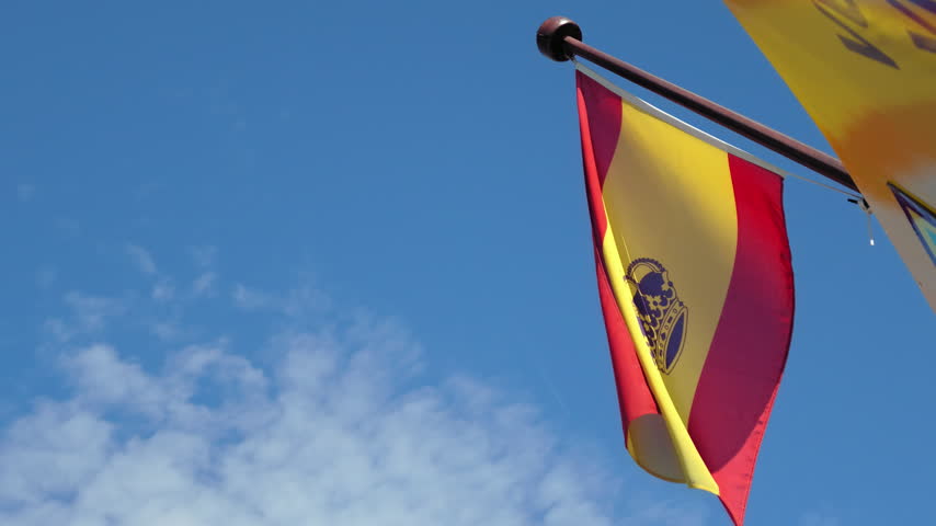 Spanish flag with a royal crown emblem waving in the wind against a clear blue sky in Maó, Menorca, Spain.