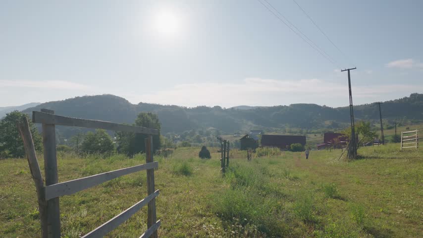 A weathered wooden fence runs down the meadow toward scattered farm buildings, hills rising behind in the haze. Filmed from a moving car, pan right with a tilt up.