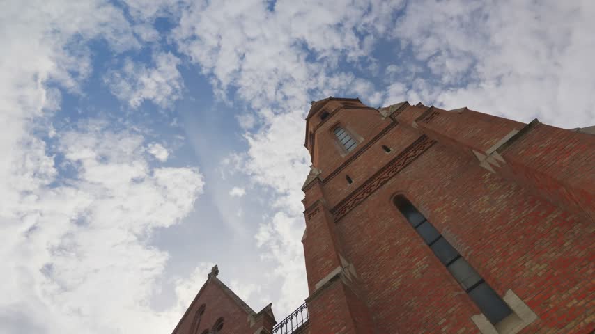 The tall brick tower and gable of the Calvinist church in Buda stand against moving cloud, a walkway linking the two. Ground-level low angle, tilt down with pan right.