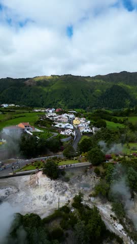 Steam rises from geothermal vents near a village with green hills in Sao Miguel