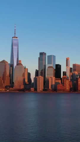 Lower Manhattan skyline glowing in golden sunset light with reflection on Hudson River calm waters during dusk, aerial footage.