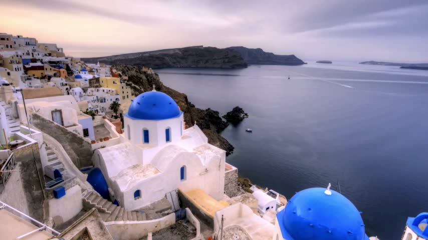 Aerial view of Oia, Santorini, Greece, showcasing its iconic whitewashed buildings, blue-domed churches, and picturesque architecture overlooking the Aegean Sea. A beautiful Mediterranean destination known for its scenic coastal views and traditional Greek charm.