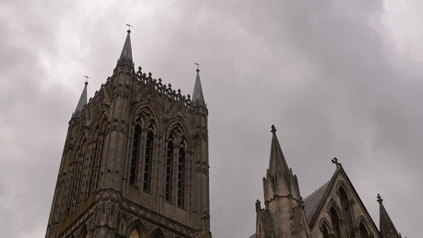 Tall cathedral spires rise into heavy grey clouds on an overcast day, framed in a static low-angle composition.