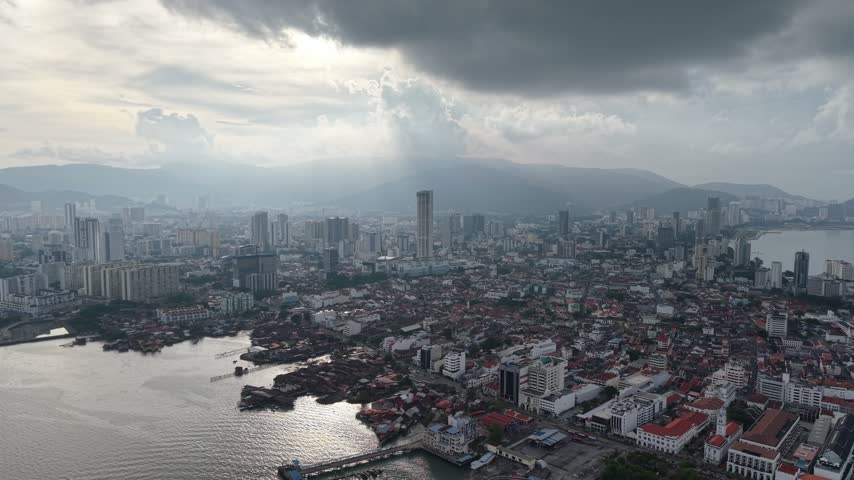 Aerial George Town cityscape along Penang waterfront, framed by dense urban architecture, mountains, dramatic clouds, with radiant sunlight.