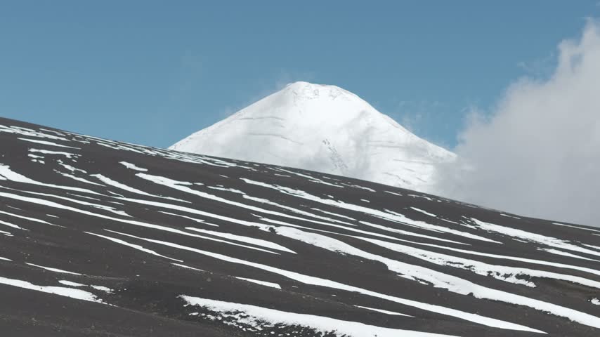 Snow-capped volcano with clouds over striped snow and ash slopes