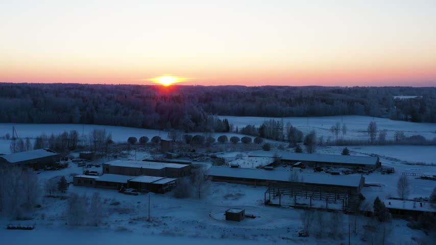 Sun setting over a winter landscape with a farm, snow-covered fields, and a frozen forest
