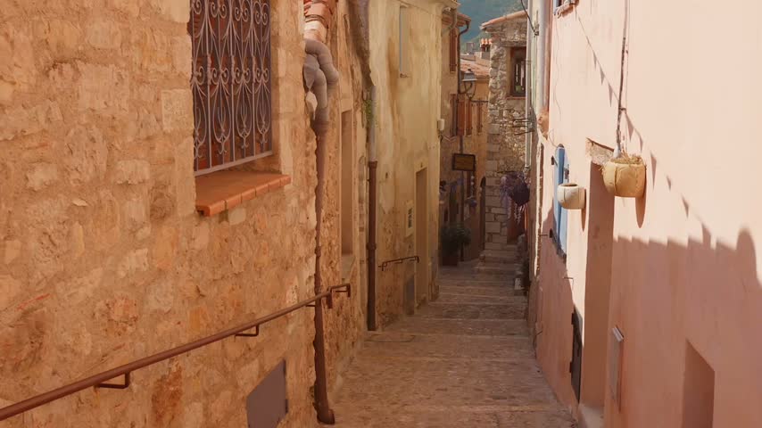 A narrow stone stairway descends between weathered masonry and pink stucco buildings, with metal handrails, hanging planters and a barred window.