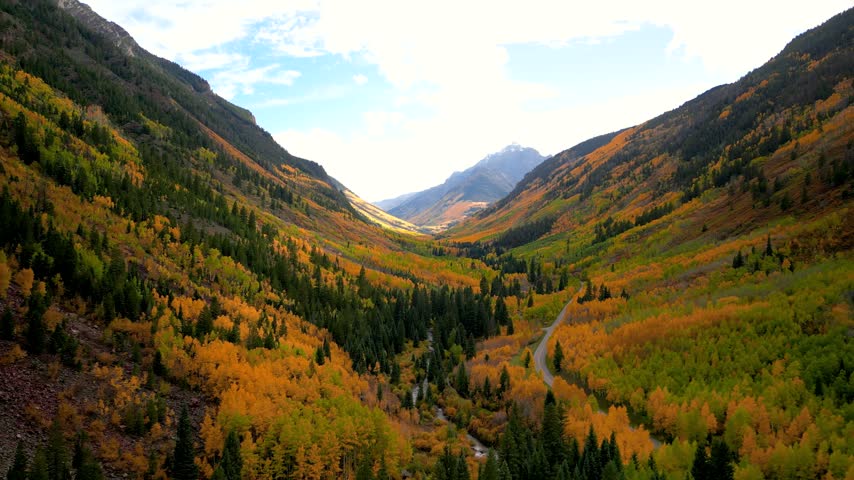 Aerial view flying through a colorful mountain valley with autumn trees and a winding road below in Colorado.