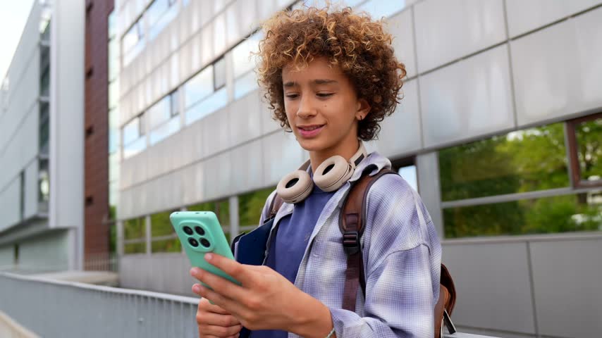 Smiling curly haired teenage boy with headphones and a backpack texting on his mobile phone, enjoying social media communication while standing in front of a modern school building