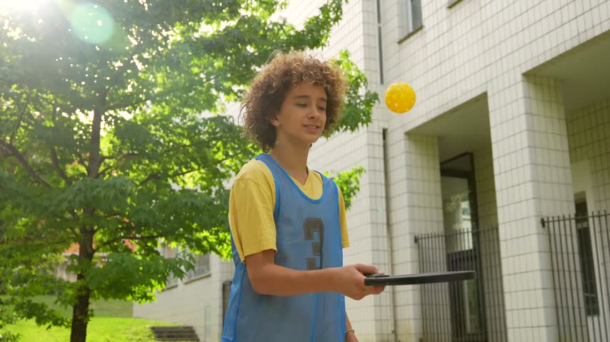 Focused teenage boy with curly hair playing pickleball with a paddle and a yellow ball, practicing his skills alone in a park with bright sunshine backlighting the scene