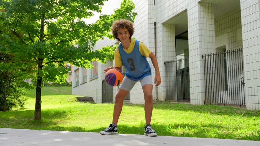 Young male student with curly hair practicing basketball, dribbling the ball with skill and confidence on a sunny day in the schoolyard, wearing his sportswear and smiling at the camera
