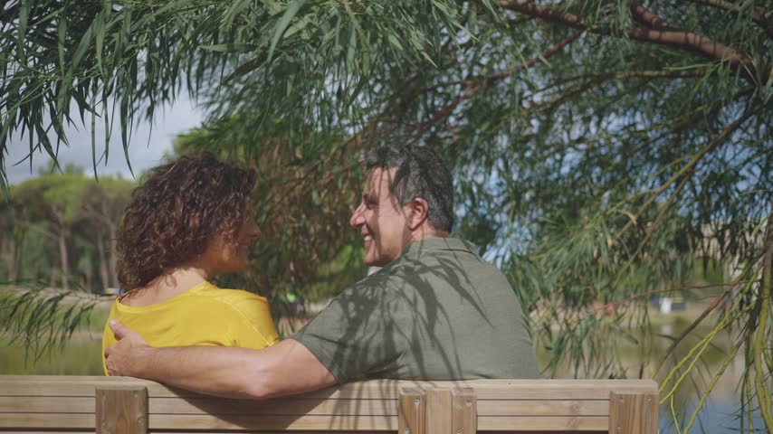 Back view of a mature couple sitting on a wooden bench by a lake under willow branches, embracing and chatting, showing love, intimacy, relaxation and togetherness.