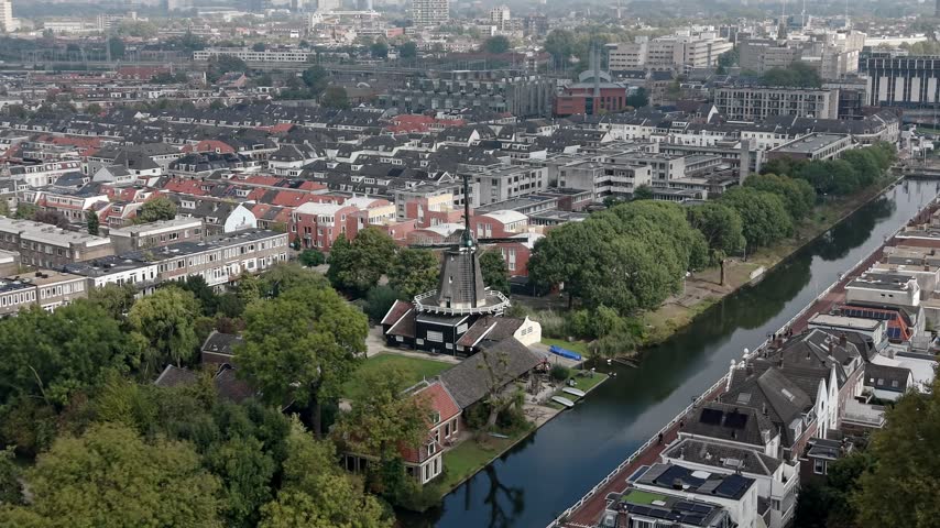Aerial view of a quiet Dutch canal bordered by leafy trees, traditional houses and dense red roof residential blocks extending across the city, 4K.