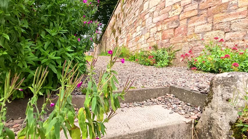 Low angle movement along rustic gravel walkway beside ancient stone wall with blooming plants and traditional town houses.