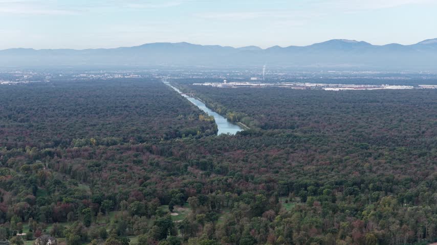 Aerial view of a canal flowing through a dense forest with distant mountains under a clear sky in Hombourg, Grand Est, France.