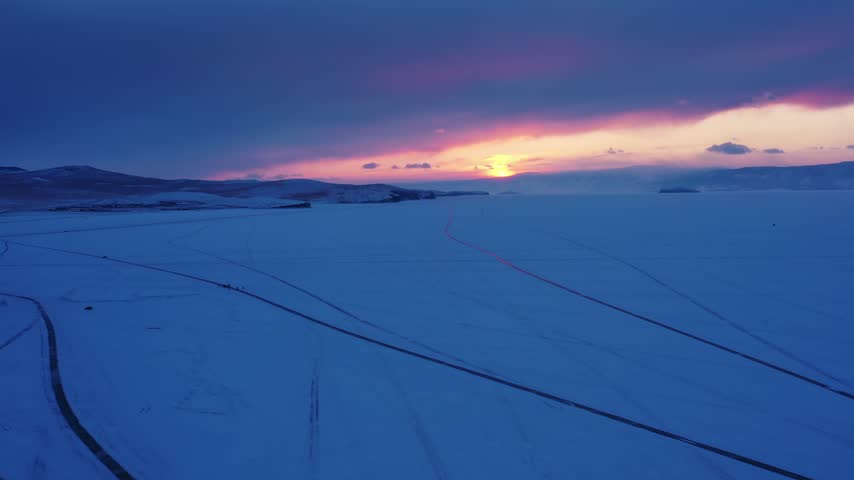 Aerial view of frozen Lake Baikal at sunset, featuring vast snow-covered ice with tracks and distant hills under a vibrant sky in Russia.