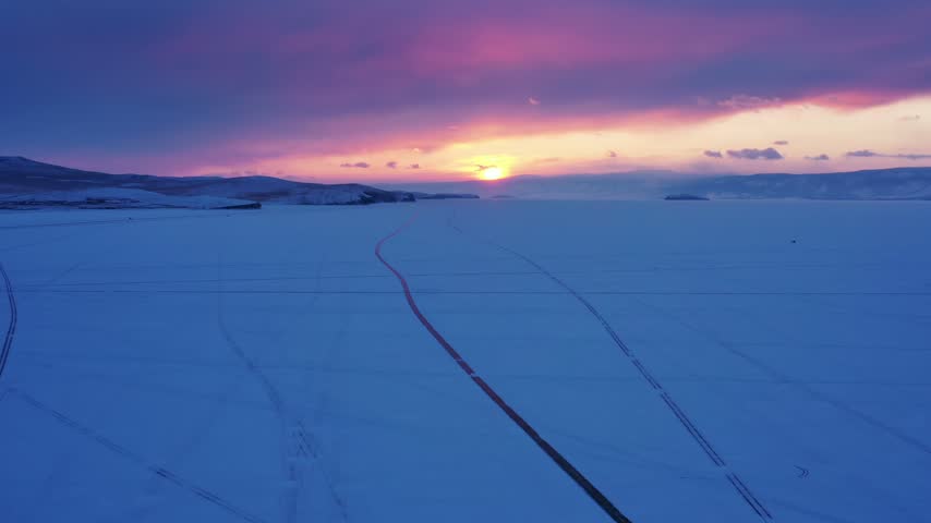 Aerial view of the frozen expanse of Lake Baikal during a vibrant sunset, featuring vast ice fields and distant snow-covered hills in Russia.