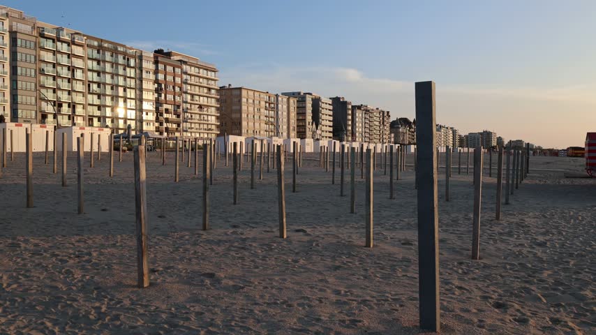 Colorful striped beach cabins line the sandy shore of De Panne, Belgium, with coastal buildings in the background.