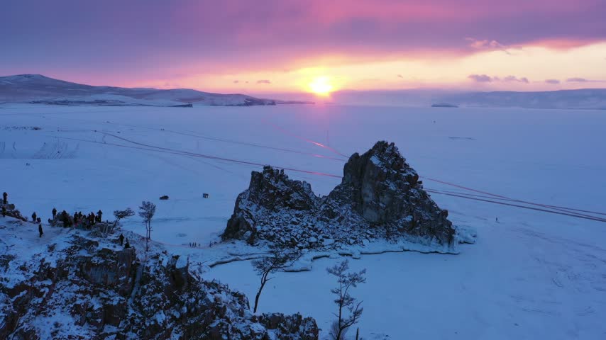 Aerial view of Shaman Rock on the frozen surface of Lake Baikal during a vibrant sunset with people gathered on the snowy shore in Russia.