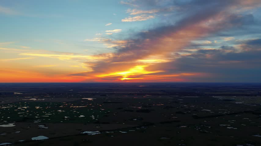 Slow forward aerial drone shot over the Canadian prairies at sunrise, with dark farmland, reflective ponds and a dramatic orange and deep blue sky as the sun breaks through clouds