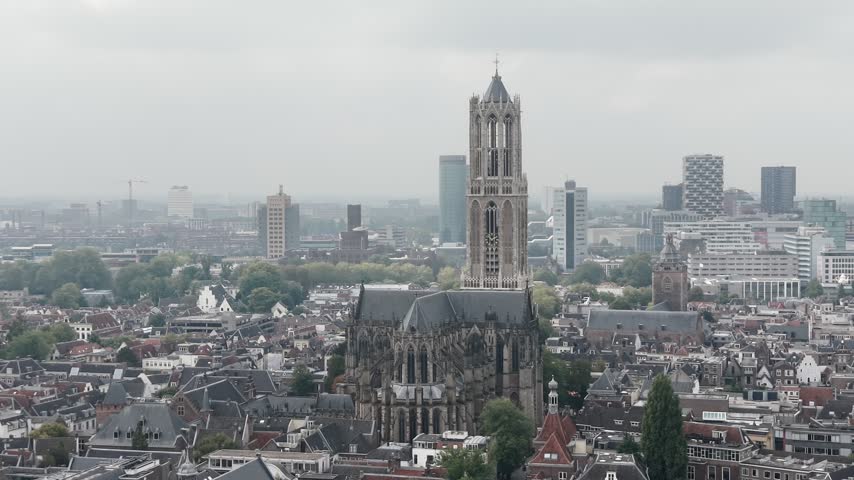 Drone view of Utrecht Dom Tower rising above historic rooftops and modern buildings in the Netherlands under a grey cloudy sky, 4K.
