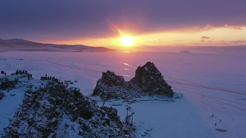 Aerial view of Lake Baikal at sunset with snow-covered rocks and people standing on the frozen surface in Russia.