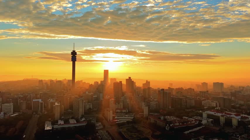 4K aerial view of Johannesburg, South Africa, at sunrise, featuring the expansive city skyline beneath a dramatic sky with fast-moving clouds, capturing the urban landscape of the country’s largest city, also known as Jozi and eGoli.