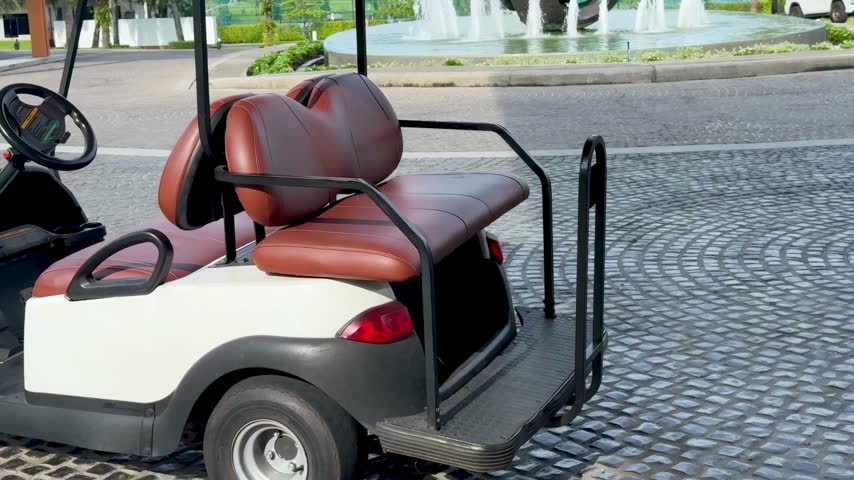 Close up side view of white golf buggy with brown seats stationary on stone pavement outdoors near luxury resort fountain.