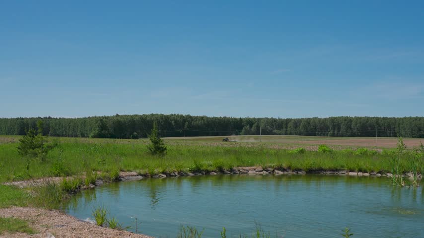 A tractor is moving across a field in the distance, near a lake